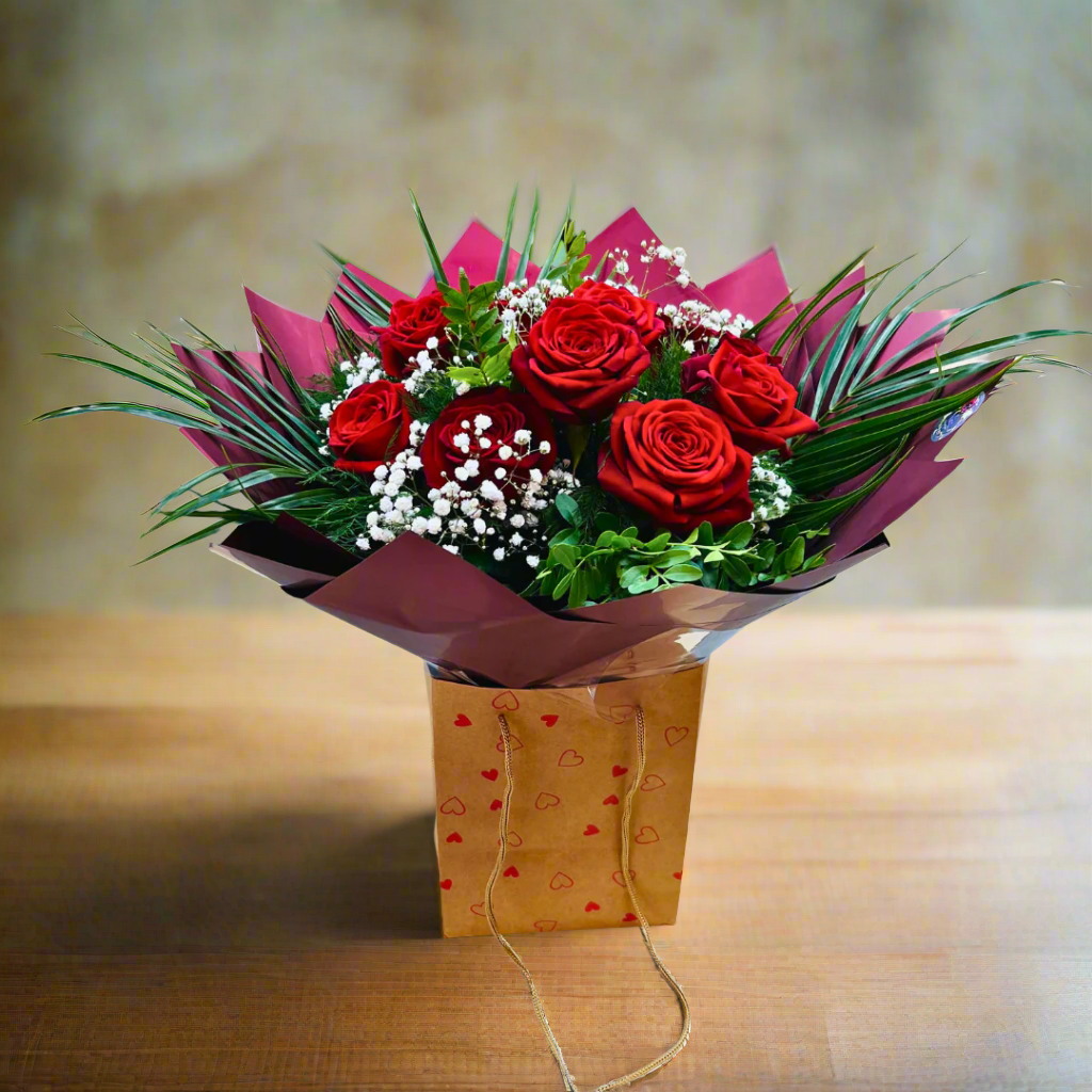 A bouquet of red roses with green leaves and white flowers, arranged in a clear vase with a red and beige paper wrap.